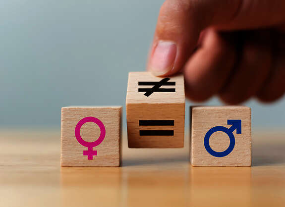 Drei kleine Holzwürfel, auf denen links ein pink-farbenes Symbol für weiblich, rechts ein blaues Symbol für männlich und dazwischen ein auf der Kippe stehender Würfen mit einem durchgestrichenen Gleichheitssymbol auf der einen und einem normalen Gleichheitszeichen auf der anderen Seite zu sehen ist