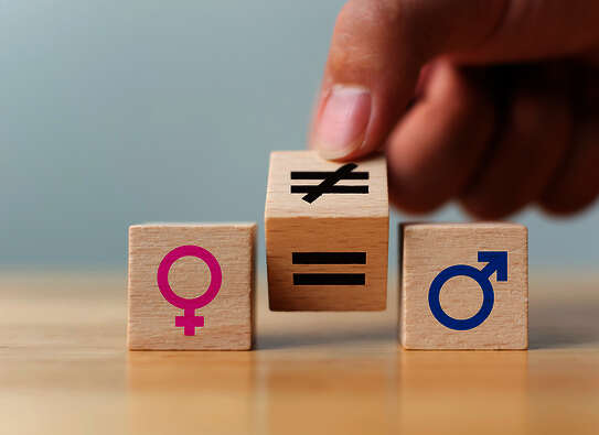 Drei kleine Holzwürfel, auf denen links ein pink-farbenes Symbol für weiblich, rechts ein blaues Symbol für männlich und dazwischen ein auf der Kippe stehender Würfen mit einem durchgestrichenen Gleichheitssymbol auf der einen und einem normalen Gleichheitszeichen auf der anderen Seite zu sehen ist